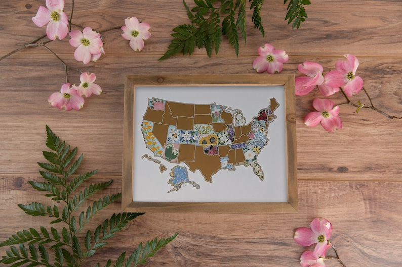 May include: A framed map of the United States with each state represented by a different floral pattern. The map is surrounded by pink dogwood blossoms and green fern leaves on a wood background.