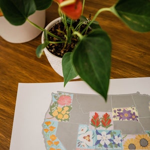 May include: A map of the United States with each state illustrated with a different flower. The states are outlined in gray and the flowers are in various colors, including red, yellow, blue, and white. The map is printed on white paper and is sitting on a wooden table.