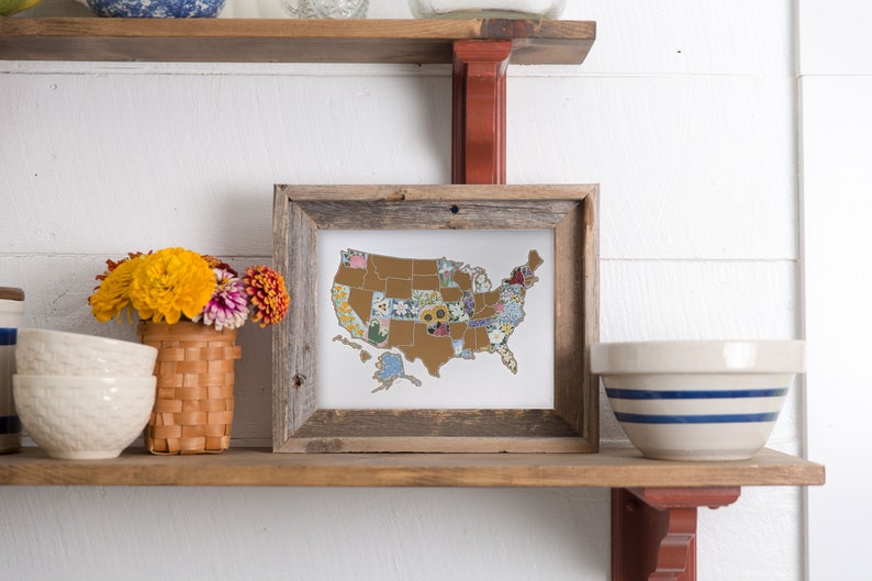 May include: A framed map of the United States with each state filled in with a different floral pattern. The map is on a wooden shelf with a vase of yellow and orange flowers and two white bowls.