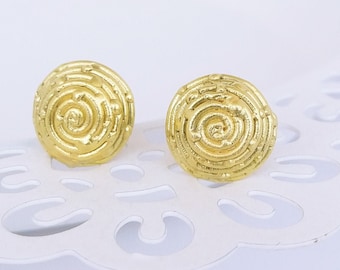 May include: A pair of gold-colored stud earrings. Each earring has a circular shape with a spiral design. The earrings are set against a white, patterned background.