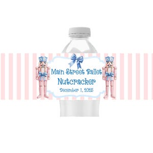 May include: A water bottle with a label featuring a pink and white striped background. The label has a light blue bow and text that reads "Main Street Ballet Nutcracker December 1, 2025". Two pink nutcracker figures are on either side.