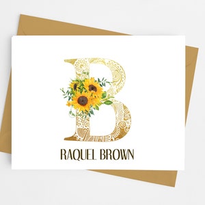 May include: A personalized stationery card featuring a gold-colored letter "B" with a decorative pattern and a bouquet of sunflowers. The name "RAQUEL BROWN" is printed below the letter in a dark brown font. The card is on a white background with a gold envelope.