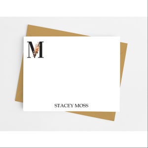 May include: A white rectangular stationery card featuring a large black letter M with a floral accent on the left side and the name STACEY MOSS printed in black at the bottom, resting on top of a tan envelope.