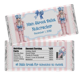 May include: A rectangular candy bar wrapper featuring a pink and white striped background. The front displays two nutcracker illustrations, a blue bow, and the text Main Street Ballet Nutcracker December 1, 2025. The back shows a nutrition facts label.