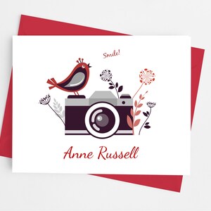 May include: Greeting card with a bird perched on a camera illustration. The card features the text "Smile!" and "Anne Russell" in a burgundy color. The card is on a red envelope.