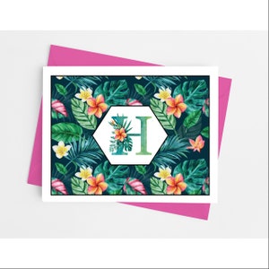 May include: A greeting card with a tropical floral design. The card features a dark blue background with green leaves and orange and yellow flowers. A white hexagon in the center displays the letter "H" in teal and green, adorned with floral accents. The card is set on a pink envelope.