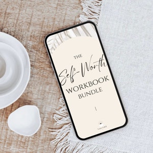 May include: A smartphone displays "The Self-Worth Workbook Bundle" in elegant script and bold text against a beige background. The phone rests on a white textured surface, with white plates and a small dish in the background.