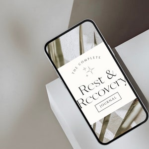 May include: A white smartphone mockup with a screen displaying a journal cover. The cover features a minimalist design with the text "The Complete Rest & Recovery Journal" and a small star graphic.
