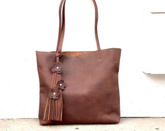 May include: A brown leather tote bag with a fringe tassel and three flower accents. The bag has a simple design and is made of high-quality leather.