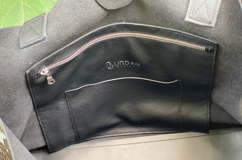 May include: The inside of a black leather tote bag with a zippered pocket and a smaller pocket. The bag has the text "Urban Artisan" printed on the inside.