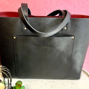 May include: Black tote bag with a textured finish and a deep red interior. The bag features a large front pocket and two handles. Gold rivets secure the handles to the bag.