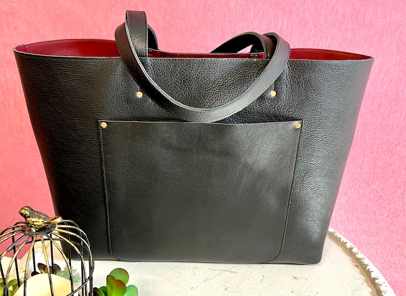 May include: Black tote bag with a textured finish and a deep red interior. The bag features a large front pocket and two handles. Gold rivets secure the handles to the bag.