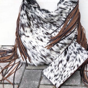 K&ouml;nnte beinhalten: Eine wei&szlig;e und schwarze Kuhfell-Handtasche mit braunen Lederfransen. Die Tasche liegt auf einer grauen Betonfl&auml;che mit einer passenden Kuhfell-Geldb&ouml;rse.