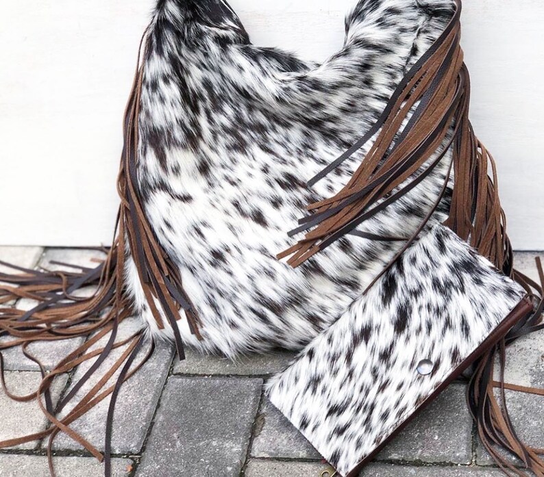 K&ouml;nnte beinhalten: Eine wei&szlig;e und schwarze Kuhfell-Handtasche mit braunen Lederfransen. Die Tasche liegt auf einer grauen Betonfl&auml;che mit einer passenden Kuhfell-Geldb&ouml;rse.