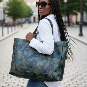May include: A large tote bag with a camouflage pattern in shades of green and blue. The bag has a rectangular shape with a front pocket and is carried over the shoulder. The person is wearing sunglasses, a white shirt, and blue jeans.