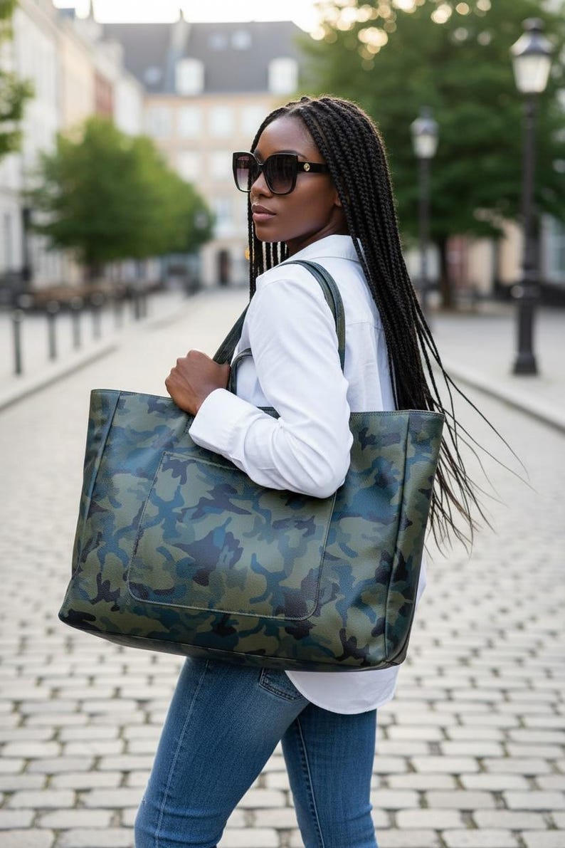 May include: A large tote bag with a camouflage pattern in shades of green and blue. The bag has a rectangular shape with a front pocket and is carried over the shoulder. The person is wearing sunglasses, a white shirt, and blue jeans.