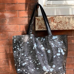 May include: A black and white cowhide tote bag with a black leather handle. The bag has a white splatter pattern.