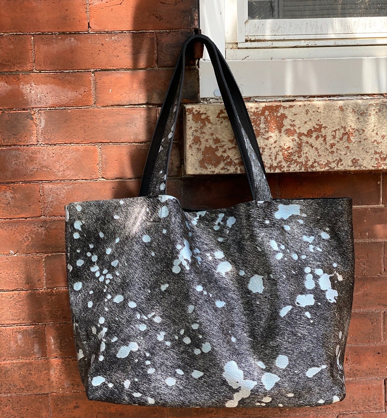 May include: A black and white cowhide tote bag with a black leather handle. The bag has a white splatter pattern.
