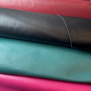 May include: A selection of rolled leather in shades of burgundy, black, teal, and magenta. The leather has a smooth surface and a subtle sheen. This material is likely intended for use in crafting, such as bags or upholstery.