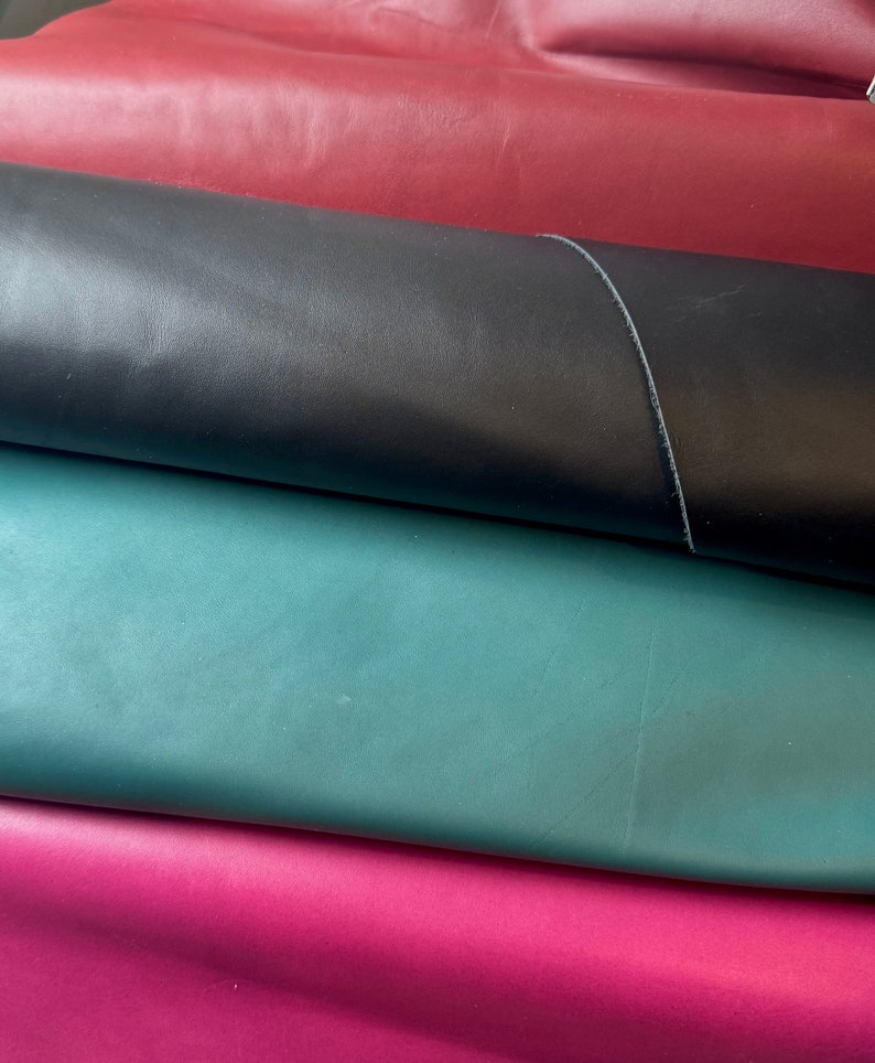 May include: A selection of rolled leather in shades of burgundy, black, teal, and magenta. The leather has a smooth surface and a subtle sheen. This material is likely intended for use in crafting, such as bags or upholstery.