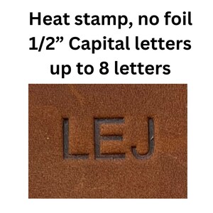 Op de afbeelding: Close-up van een bruin lederen oppervlak met de letters "LEJ" in het materiaal gebrand. De tekst boven de letters luidt "Heat stamp, no foil 1/2" hoofdletters tot 8 letters."