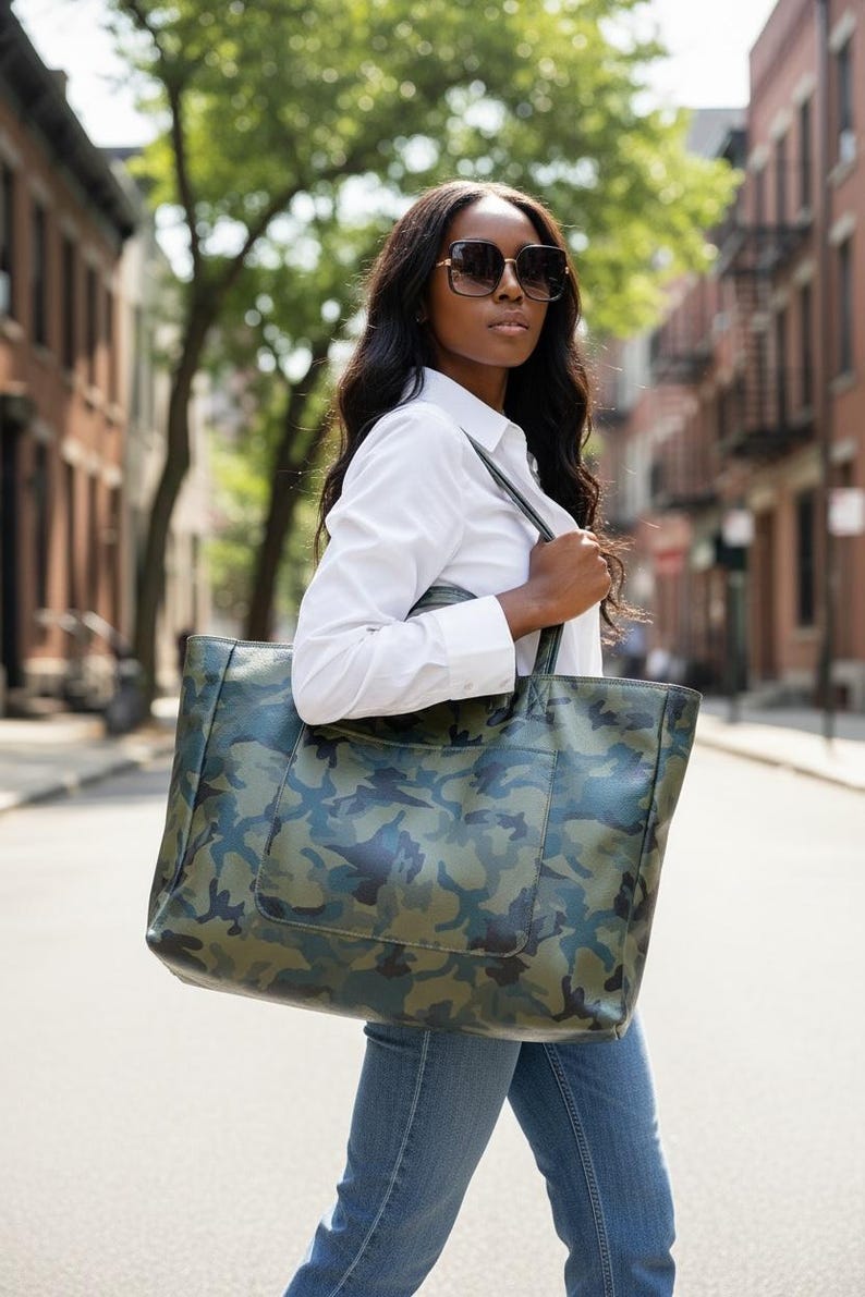 May include: A large tote bag with a camouflage pattern in shades of green, blue, and brown. The bag is carried over the shoulder. The person is wearing sunglasses, a white shirt, and blue jeans.