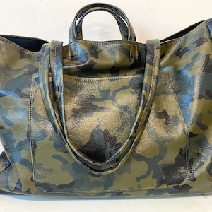 May include: A large tote bag with a camouflage pattern in shades of green, black, and brown. The bag has two short handles and a shoulder strap. The bag is made of a textured material.