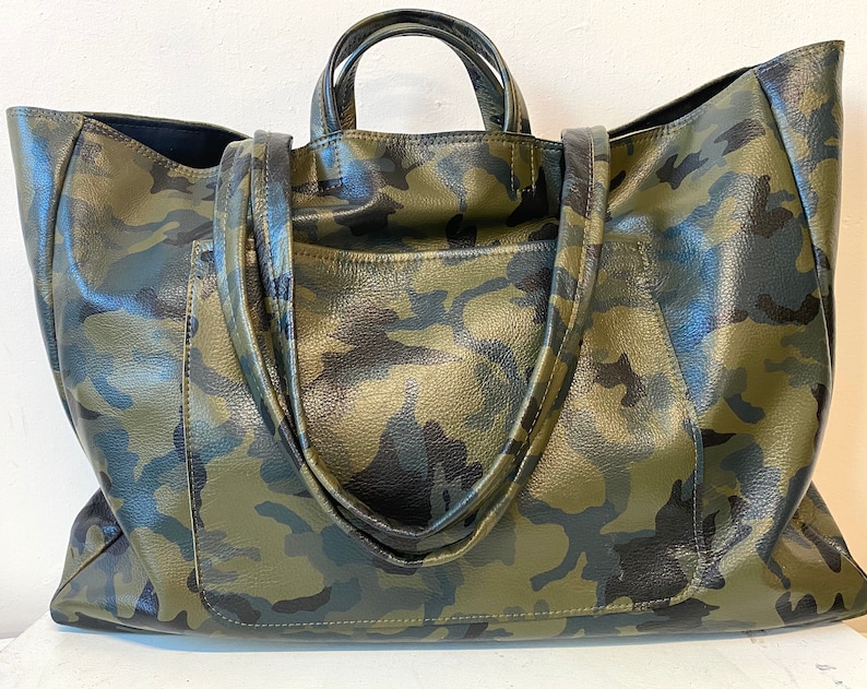 May include: A large tote bag with a camouflage pattern in shades of green, black, and brown. The bag has two short handles and a shoulder strap. The bag is made of a textured material.