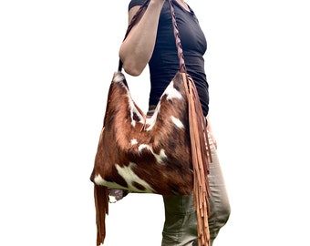 May include: A brown and white cowhide shoulder bag with long leather fringe. The bag has a braided leather strap and is being carried over the shoulder. The bag's texture and color variations are prominent.