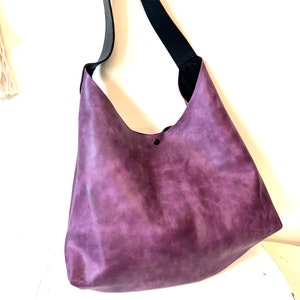 Puede incluir: Un bolso tote de cuero morado con una correa negra. El bolso tiene un aspecto desgastado y un solo bot&oacute;n negro en la parte delantera.