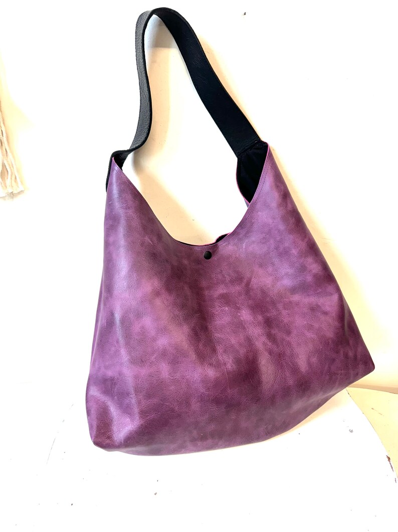 Puede incluir: Un bolso tote de cuero morado con una correa negra. El bolso tiene un aspecto desgastado y un solo bot&oacute;n negro en la parte delantera.