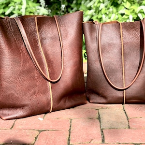 May include: Two brown leather tote bags with tan stitching. The larger bag is on the left and the smaller bag is on the right.