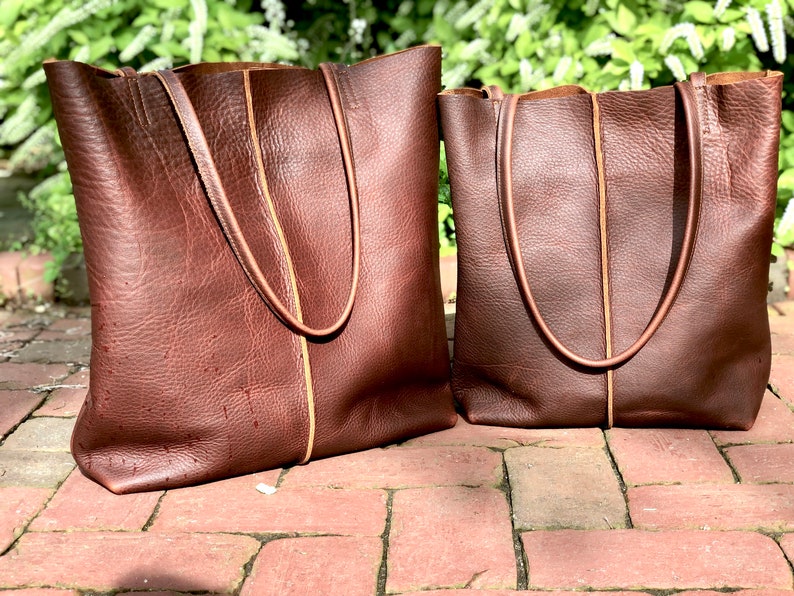 May include: Two brown leather tote bags with tan stitching. The larger bag is on the left and the smaller bag is on the right.