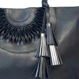 Op de afbeelding: Zwarte leren tote bag met een getextureerde afwerking. De tas heeft een decoratief halfrond ontwerp en bungelende kwastjes in zwart en zilver. De tas heeft een handvat en is geschikt voor dagelijks gebruik.