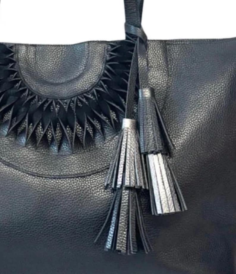 Op de afbeelding: Zwarte leren tote bag met een getextureerde afwerking. De tas heeft een decoratief halfrond ontwerp en bungelende kwastjes in zwart en zilver. De tas heeft een handvat en is geschikt voor dagelijks gebruik.