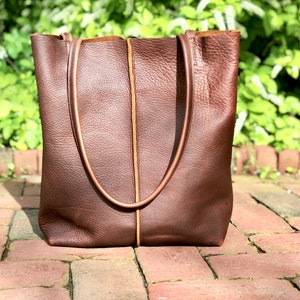 May include: A brown leather tote bag with a tan leather trim. The bag has two long handles and a wide opening.