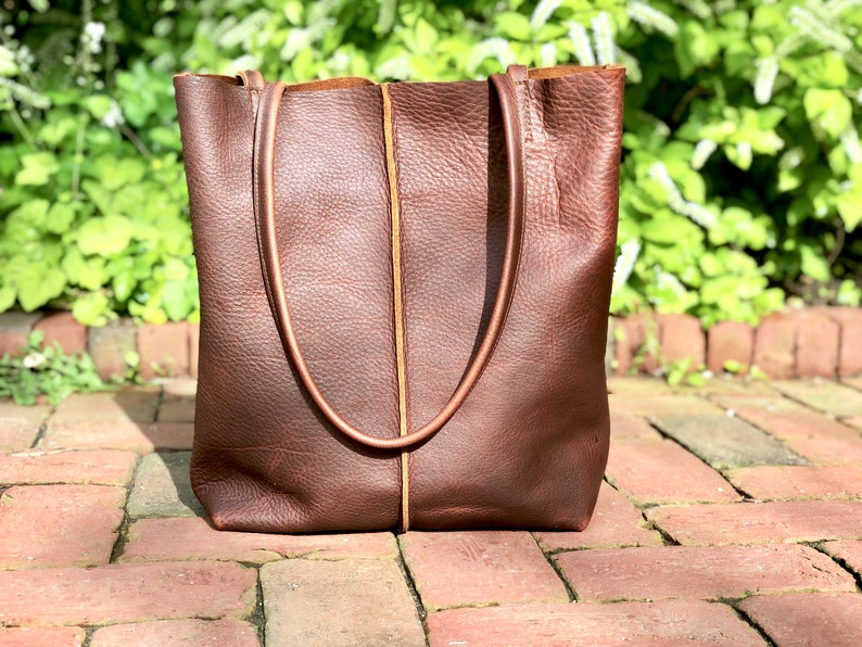 May include: A brown leather tote bag with a tan leather trim. The bag has two long handles and a wide opening.