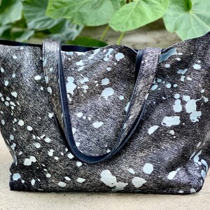 May include: A black and white cowhide tote bag with black leather handles. The bag has a white splatter pattern.