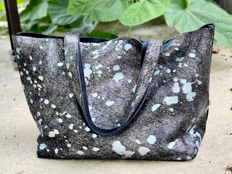 May include: A black and white cowhide tote bag with black leather handles. The bag has a white splatter pattern.