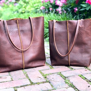 May include: Two brown leather tote bags with tan stitching. The larger bag is on the left and the smaller bag is on the right. Both bags have a rectangular shape and two handles.