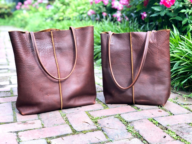 May include: Two brown leather tote bags with tan stitching. The larger bag is on the left and the smaller bag is on the right. Both bags have a rectangular shape and two handles.