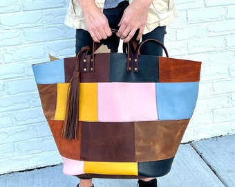 May include: A large patchwork tote bag made of different colored leather. The bag has a brown tassel and is held by a person wearing a white jacket and black pants.