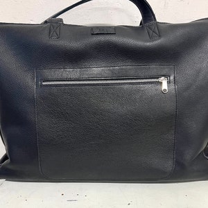 Op de afbeelding: Een zwarte leren tote bag met een zilveren ritsvak aan de voorkant. De tas heeft een handvat aan de bovenkant en een klein rechthoekig label met de letters "MEL". De tas is gemaakt van een textuur.