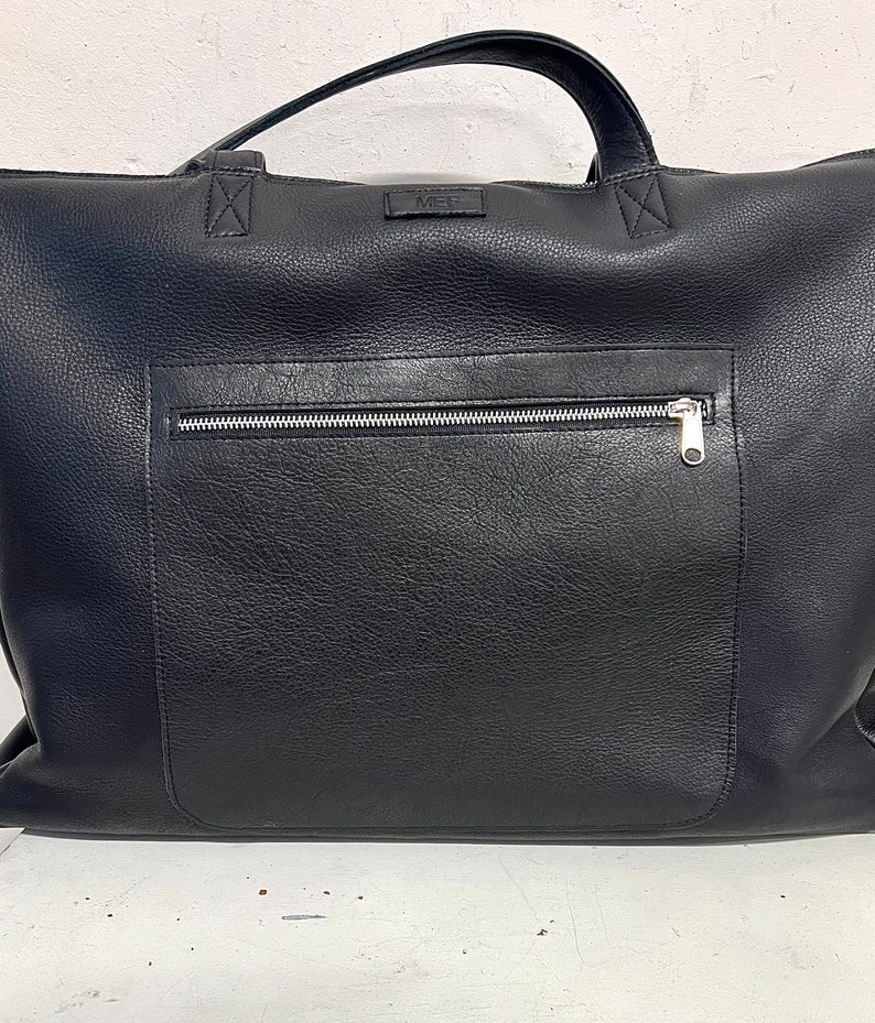 Op de afbeelding: Een zwarte leren tote bag met een zilveren ritsvak aan de voorkant. De tas heeft een handvat aan de bovenkant en een klein rechthoekig label met de letters "MEL". De tas is gemaakt van een textuur.