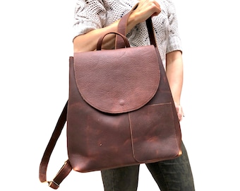 May include: A brown leather backpack with a flap closure and a front pocket. The backpack has adjustable shoulder straps and a top handle. The leather has a textured appearance. The bag is designed for carrying items.