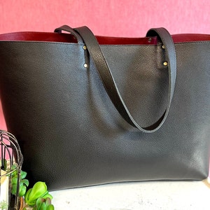 May include: A large, dark grey tote bag with a textured exterior and a deep red interior. The bag has two long handles and is accented with gold-coloured rivets. The bag is displayed against a pink background.