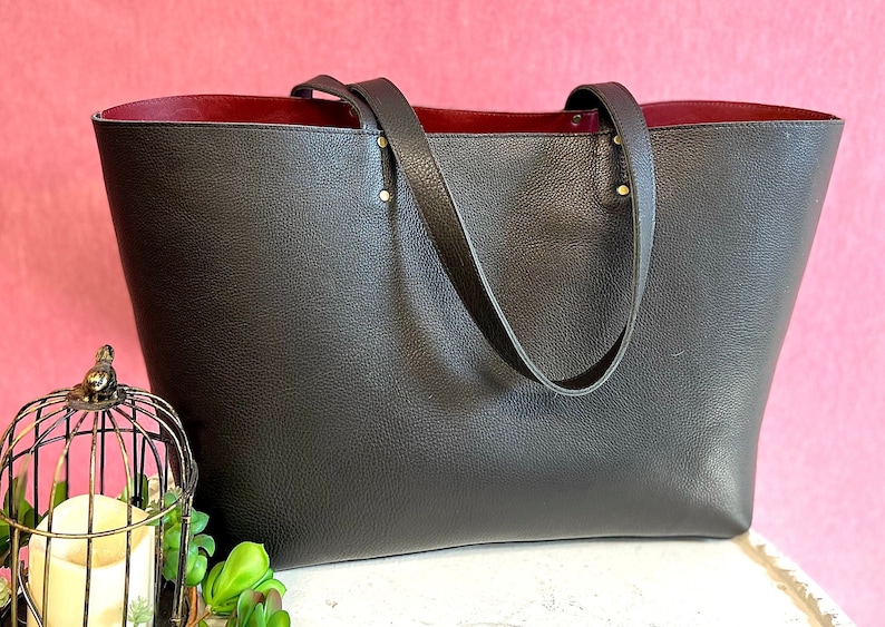 May include: A large, dark grey tote bag with a textured exterior and a deep red interior. The bag has two long handles and is accented with gold-coloured rivets. The bag is displayed against a pink background.