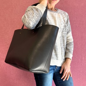May include: A large, black tote bag with a textured finish and long handles. The bag is held by a person wearing a grey and white sweatshirt and blue jeans. The background is a solid pink colour.