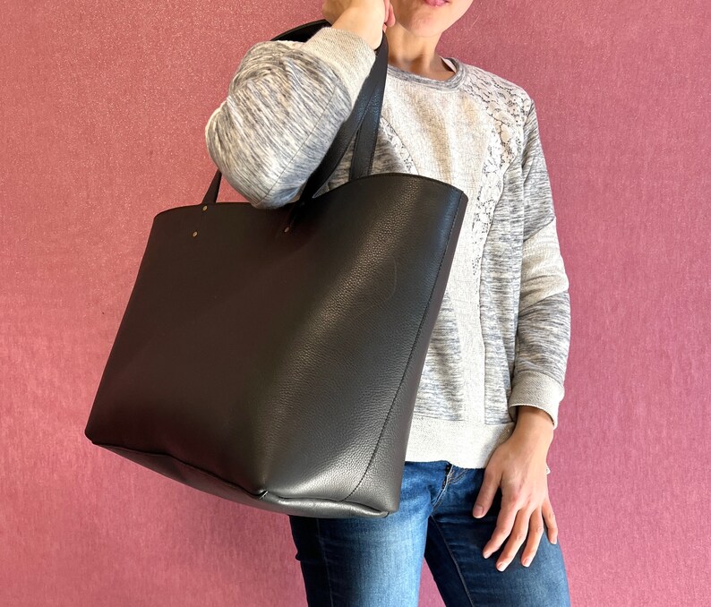 May include: A large, black tote bag with a textured finish and long handles. The bag is held by a person wearing a grey and white sweatshirt and blue jeans. The background is a solid pink colour.