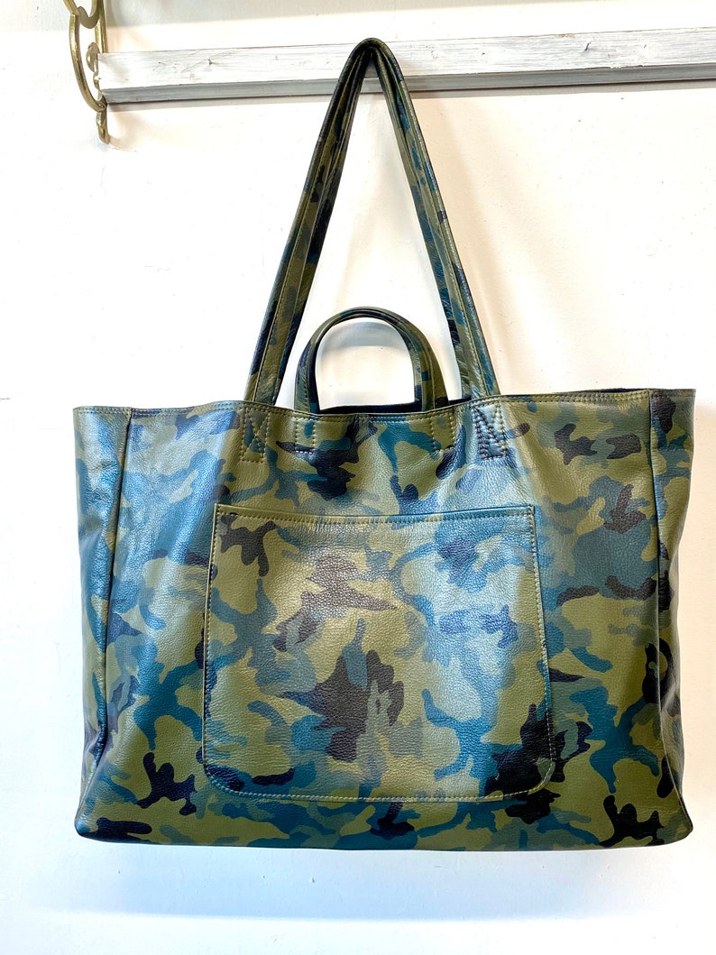 May include: A large tote bag with a camouflage pattern in shades of green, blue, and black. The bag has a front pocket and two long handles. The bag is hanging against a white background.